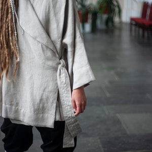May include: A light gray linen wrap top with a long, flowing sleeve and a tie belt. The top is worn over black pants and white boots.