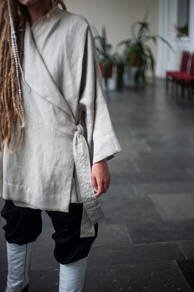 May include: A light gray linen wrap top with a long, flowing sleeve and a tie belt. The top is worn over black pants and white boots.