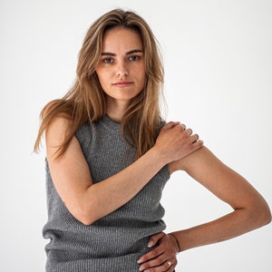 Op de afbeelding: Een vrouw draagt een grijs, mouwloos, geribbeld gebreid topje en een donkergroene corduroy broek. Ze staat voor een witte achtergrond.
