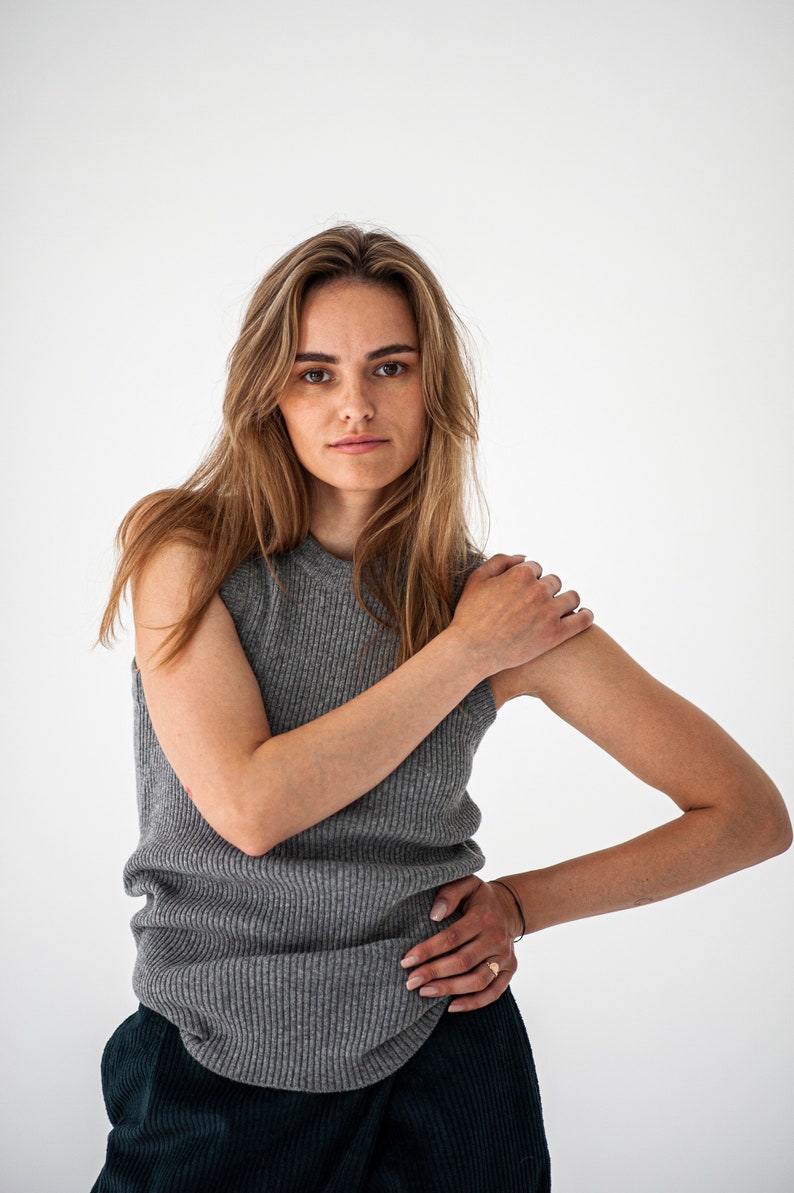 Op de afbeelding: Een vrouw draagt een grijs, mouwloos, geribbeld gebreid topje en een donkergroene corduroy broek. Ze staat voor een witte achtergrond.