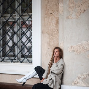 May include: A woman with long dreadlocks sits on a wooden bench wearing a light gray linen jacket, black pants, and white knee-high boots. She is leaning against a white wall with a window behind her.