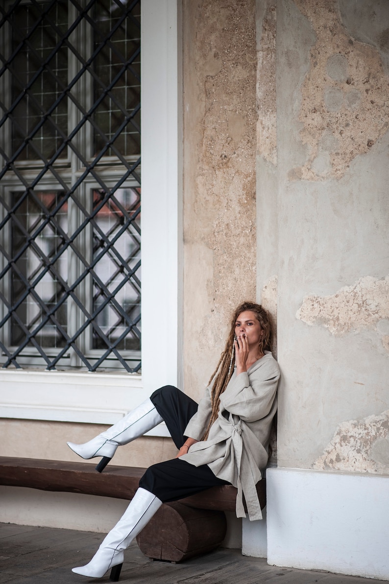 May include: A woman with long dreadlocks sits on a wooden bench wearing a light gray linen jacket, black pants, and white knee-high boots. She is leaning against a white wall with a window behind her.