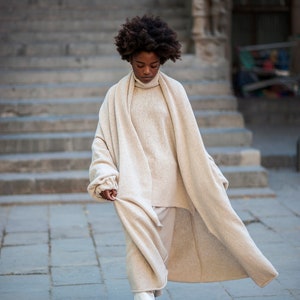 May include: A woman wearing a long, cream-colored knitted cardigan over a cream turtleneck sweater and white pants. She is wearing white boots and has curly dark hair.