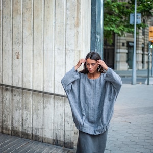 May include: A woman wearing a grey oversized sweater with a herringbone pattern and a long grey skirt. She is standing in front of a concrete wall.