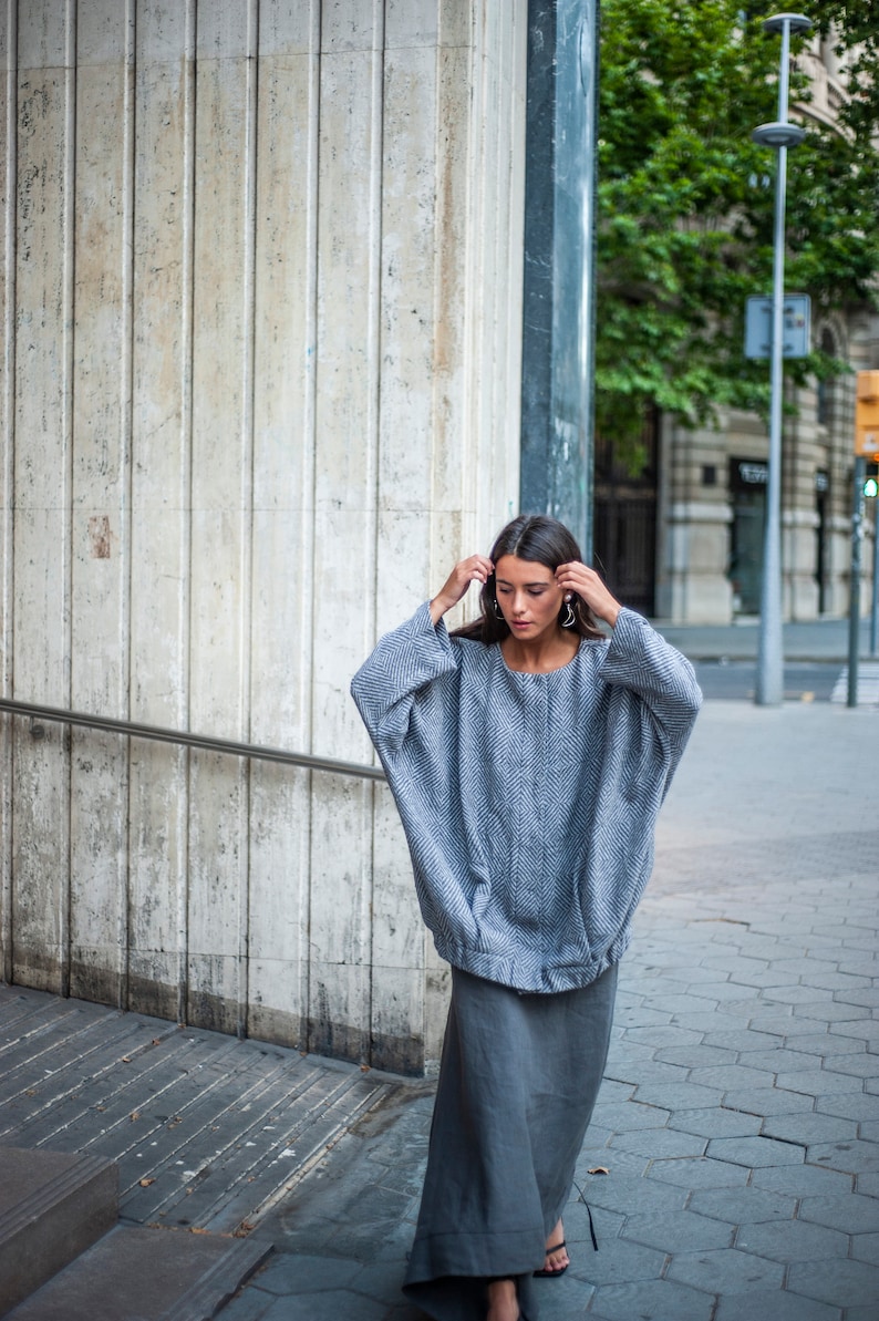 May include: A woman wearing a grey oversized sweater with a herringbone pattern and a long grey skirt. She is standing in front of a concrete wall.