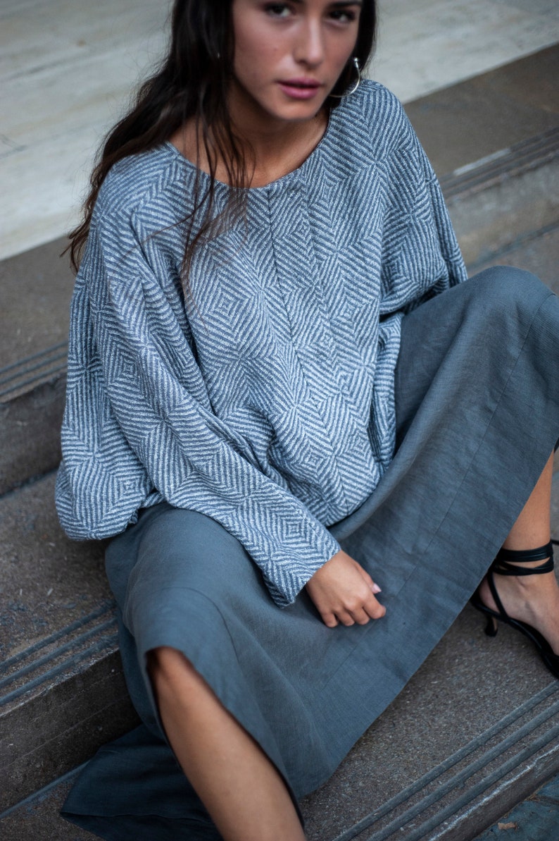May include: A woman wearing a grey and white patterned blouse with long sleeves and a grey skirt. She is sitting on a set of stairs.