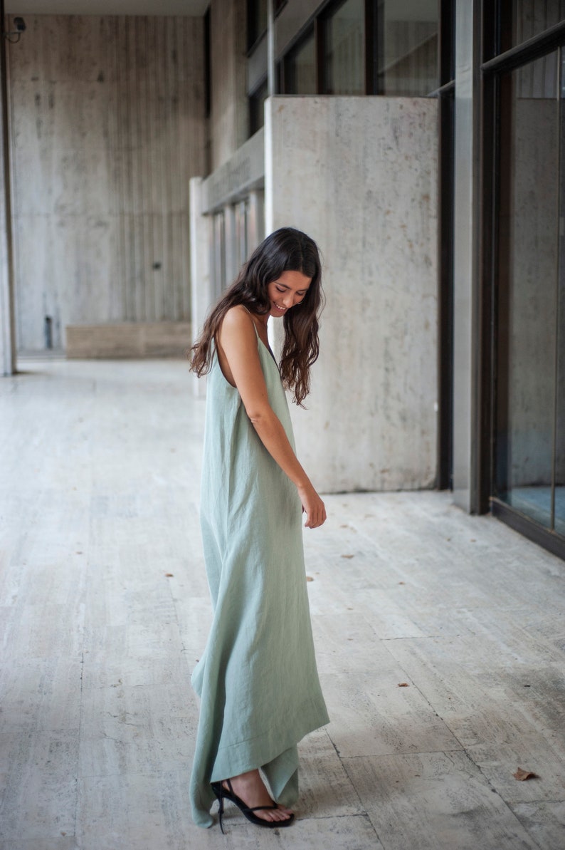 May include: A woman wearing a sage green linen maxi dress with a simple, sleeveless design. The dress has a flowing silhouette and is slightly gathered at the waist. She is standing on a light-colored tiled floor.