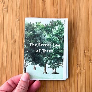 May include: A small rectangular booklet with a white border featuring a watercolour illustration of green trees and the text The Secret Life of Trees printed in white. The cover art depicts a forest scene with dark and light green foliage.