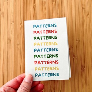 May include: A white rectangular greeting card featuring the word PATTERNS printed nine times in a vertical column. Each instance of the word is written in a different color, including blue, red, green, and yellow, using a rounded sans-serif font.
