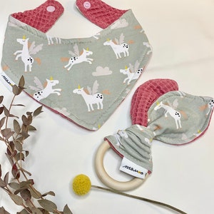 May include: A set of two baby items featuring a unicorn design. A green and pink bib with a waffle texture and a wooden teether with a pink and green fabric loop.