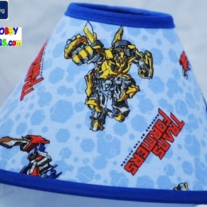 May include: A blue and yellow Transformers-themed baby neck support. The neck support is shaped like a cone and has a blue trim. The fabric features images of Optimus Prime and Bumblebee, along with the word "Transformers".