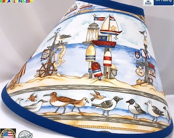May include: A blue and white lampshade with a nautical theme. The lampshade features a beach scene with a lighthouse, sailboat, buoys, and seagulls. The lampshade has a blue trim.