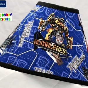 May include: A blue and black lampshade with a Transformers theme. The lampshade features images of Optimus Prime, Bumblebee, and other Transformers characters. The text "Transformers" is printed on the lampshade.