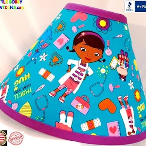 May include: A turquoise lamp shade featuring a cartoon doctor character, bandages, stethoscopes, and other medical-themed illustrations. The shade has a purple trim at the top and bottom. The text "THAT'LL cost you a HUG" is also visible.