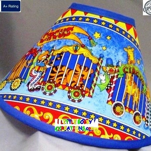 May include: A blue and red lampshade with a circus theme. The lampshade features a tiger in a cage, a clown, and a giraffe. The text "Circus" is printed on the lampshade. The text "I Little Bobby's Creations.com" is printed on the bottom of the lampshade.