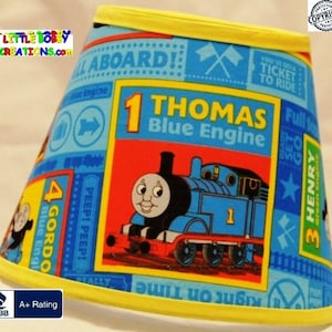 May include: A blue and yellow lampshade with a Thomas the Tank Engine design. The lampshade features a large image of Thomas the Tank Engine, along with the text "1 Thomas Blue Engine" and other details from the Thomas & Friends franchise.