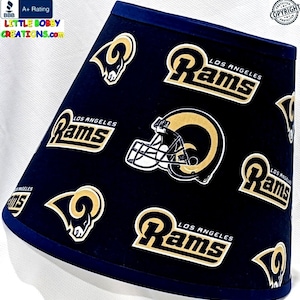 May include: A blue lampshade with a repeating pattern of the Los Angeles Rams logo. The logo features a gold ram's head with horns and the words "Los Angeles Rams" in gold lettering.