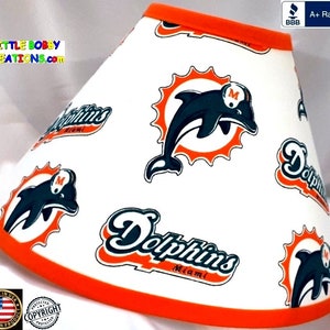 May include: White lampshade with an orange trim featuring a repeating pattern of the Miami Dolphins logo. The logo features a dolphin jumping out of a sunburst with the word "Dolphins" and "Miami" written below.