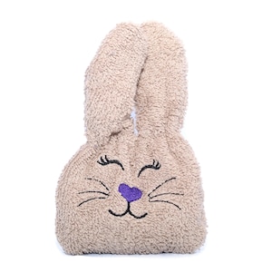 Puede incluir: Una bolsa de peluche beige con forma de conejo con ojos bordados, una nariz morada y bigotes.