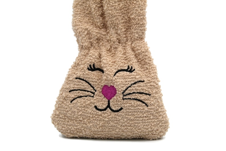 Puede incluir: Una bolsa de peluche marr&oacute;n con forma de conejo con un coraz&oacute;n rosa y bigotes y pesta&ntilde;as bordados en negro.