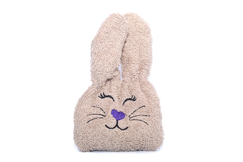 Puede incluir: Una bolsa de peluche beige con forma de conejo con ojos bordados, una nariz morada y bigotes.