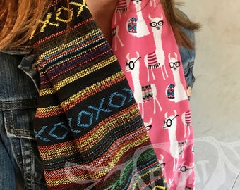 May include: Two infinity scarves. One is black with colorful horizontal stripes and the word "XOXO". The other is pink with a pattern of white llamas wearing glasses. The scarves are worn over a denim jacket.