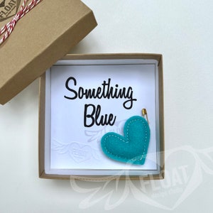 Könnte beinhalten: Türkisfarbene Filzherz-Anstecknadel mit weißen Nähten und einer goldenen Sicherheitsnadel, präsentiert in einer braunen Geschenkbox. Die Box enthält eine weiße Karte mit dem Schriftzug "Something Blue".