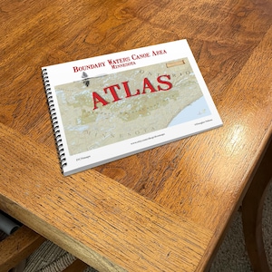 May include: A spiral-bound atlas titled "Boundary Waters Canoe Area Minnesota" with a map of the region. The word "ATLAS" is in large red letters. The atlas is on a wooden table.