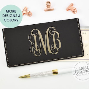 May include: A black leather checkbook cover with a gold monogram "MB" and a stitched edge. The cover is lying on a white surface with a pen and a checkbook.