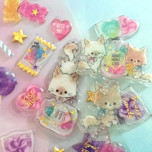 Puede incluir: Una hoja de pegatinas coloridas y brillantes con gatos, corazones, caramelos y estrellas. Las pegatinas están hechas de plástico transparente con un efecto 3D. El texto en las pegatinas incluye "Kiss Hug", "Sweet Candy", "Mero Mero Cats" y "Kiss Me".
