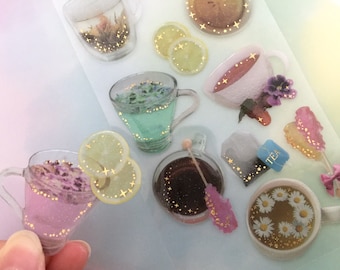 Könnte beinhalten: Dekorative Aufkleber mit Tee-Motiven. Die Aufkleber zeigen Teetassen mit floralen Aufgüssen, Zitronenscheiben und einen Teebeutel. Goldglitzer und Sternchen-Akzente sind enthalten. Das Thema ist Tee und Getränke.