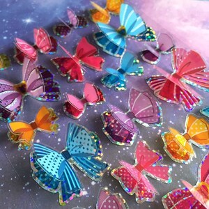 以下が含まれることがあります： ピンク、ブルー、オレンジ、パープルのアクセントが付いた虹色のホログラフィックな蝶のステッカーのシート。ステッカーは紫色の背景にランダムなパターンで配置されています。