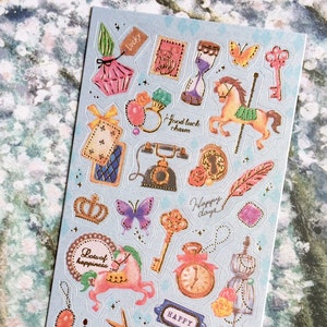 May include: A sheet of colorful stickers with a variety of designs, including butterflies, a carousel horse, a birdcage, a crown, a key, a clock, a bottle of perfume, a telephone, a scissors, a playing card, and a feather. The stickers have gold accents and are on a light blue background.