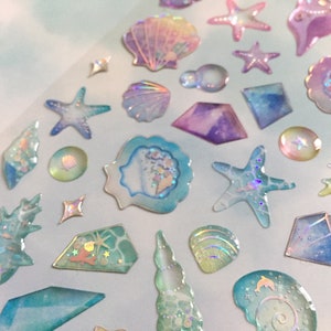 Pode incluir: Conjunto de adesivos iridescentes com conchas, estrelas do mar e outros designs com tema oceânico. Os adesivos são translúcidos e têm um acabamento holográfico.