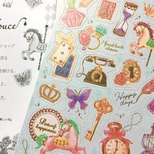 May include: A sheet of decorative stickers featuring a variety of whimsical designs, including a carousel horse, a crown, a key, a feather, a clock, a butterfly, a telephone, a bottle of perfume, and a unicorn. The stickers are printed on a light blue background with gold accents.