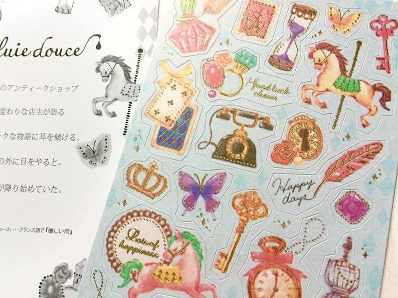 May include: A sheet of decorative stickers featuring a variety of whimsical designs, including a carousel horse, a crown, a key, a feather, a clock, a butterfly, a telephone, a bottle of perfume, and a unicorn. The stickers are printed on a light blue background with gold accents.
