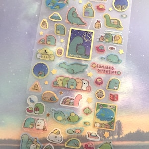 Puede incluir: Una hoja de pegatinas transparentes con varias ilustraciones de personajes de dibujos animados lindos. Los personajes son principalmente de color verde pastel y azul, con algunos acentos amarillos y rosados. Los personajes son de diferentes tamaños y formas, y algunos sostienen objetos como libros, estrellas y peces. Las pegatinas están dispuestas en un patrón de cuadrícula sobre un fondo blanco. El texto "Chase the stars" está en la parte superior de la hoja, y el texto "Sumikko Gurashi" está en la parte inferior.