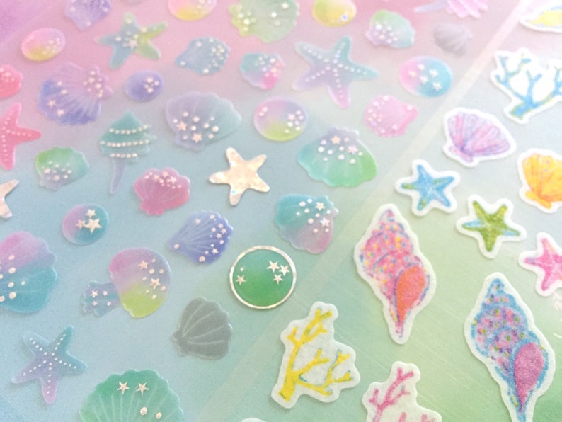 May include: A sheet of iridescent stickers featuring various sea-themed designs, including seashells, starfish, coral, and a Christmas tree. The stickers are translucent and have a glittery finish.
