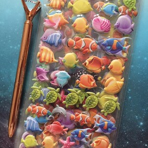 Puede incluir: Una bolsa de pl&aacute;stico transparente llena de pegatinas de espuma 3D de colores de criaturas marinas, incluyendo peces, tortugas y estrellas de mar. Las pegatinas est&aacute;n dispuestas en filas y columnas, y hay una variedad de colores y dise&ntilde;os. Un bol&iacute;grafo dorado con una parte superior en forma de diamante est&aacute; al lado de la bolsa.