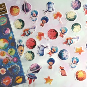 Puede incluir: Una hoja de pegatinas coloridas con tem&aacute;tica espacial que representan planetas, estrellas, cohetes y animales con cascos de astronauta. Las pegatinas est&aacute;n sobre un fondo blanco y tienen un aspecto de acuarela. La hoja est&aacute; etiquetada como "Nekoni glitter&elegant".