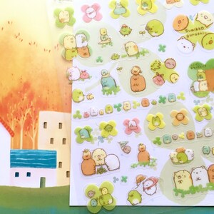 May include: A sheet of clear stickers featuring adorable cartoon characters. The characters are mostly white and beige with brown, pink, and yellow accents. The stickers are on a white background with green clover shapes. The text "Sumikko gurashi" is printed in black on a yellow sticker.