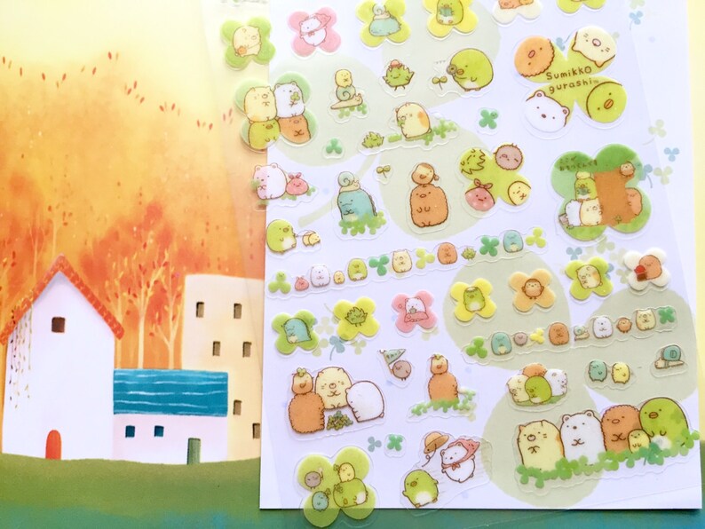 May include: A sheet of clear stickers featuring adorable cartoon characters. The characters are mostly white and beige with brown, pink, and yellow accents. The stickers are on a white background with green clover shapes. The text "Sumikko gurashi" is printed in black on a yellow sticker.