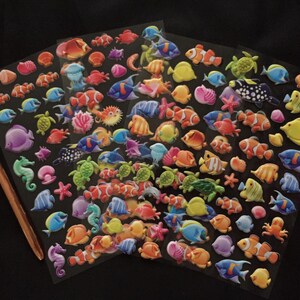 Puede incluir: Una hoja de pegatinas coloridas en 3D, autoadhesivas, que representan diversas criaturas marinas, incluyendo peces, caballitos de mar, estrellas de mar y conchas. Las pegatinas est&aacute;n en una hoja de pl&aacute;stico transparente.