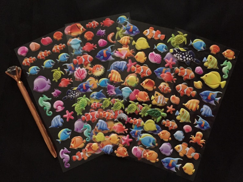 Puede incluir: Una hoja de pegatinas coloridas en 3D, autoadhesivas, que representan diversas criaturas marinas, incluyendo peces, caballitos de mar, estrellas de mar y conchas. Las pegatinas est&aacute;n en una hoja de pl&aacute;stico transparente.