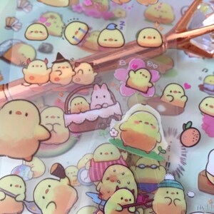 以下が含まれることがあります： 様々なポーズと衣装を着た、黄色い漫画のひよこのデザインが施された、透明な粘着ステッカーのシート。ステッカーは白い背景にあり、帽子をかぶったひよこ、花を持っているひよこ、サーフボードに乗っているひよこなど、様々なデザインが含まれています。