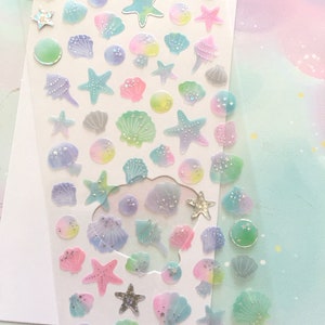 May include: A sheet of iridescent stickers featuring seashells and starfish in various pastel colors. The stickers are on a clear background and have a glittery finish. The sheet is labeled "YT-HT012".