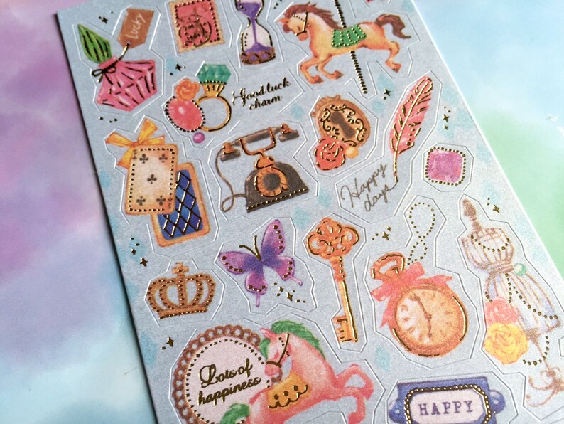 May include: A sheet of decorative stickers with a blue background and gold accents. The stickers feature various images, including a carousel horse, a butterfly, a crown, a key, a clock, a feather, a telephone, a perfume bottle, a playing card, a ring, a cage, and a tag that says "lucky".
