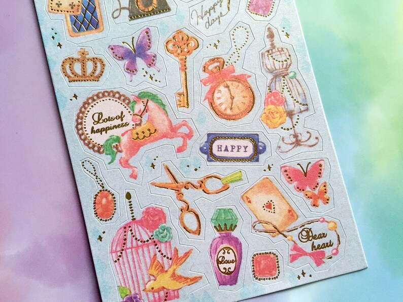 May include: A sheet of colorful stickers with a blue background. The stickers feature various images, including a crown, a butterfly, a horse, a birdcage, a bird, a clock, a key, a pair of scissors, a bottle of perfume, a playing card, a heart, and a necklace. The stickers also include the words "Lots of happiness", "Happy", and "Dear heart".