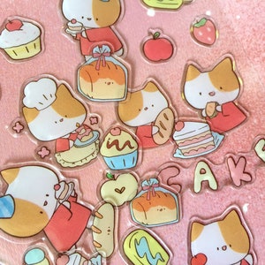 K&ouml;nnte beinhalten: Ein Set bunter Aufkleber mit Comic-Katzen mit verschiedenen Gesichtsausdr&uuml;cken und verschiedenen Lebensmitteln, darunter Brot, Kuchen und Cupcakes. Die Aufkleber befinden sich auf einem rosa Hintergrund und haben eine gl&auml;nzende Oberfl&auml;che.