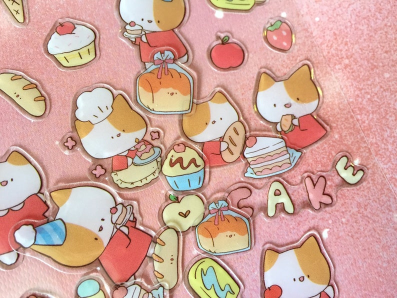 K&ouml;nnte beinhalten: Ein Set bunter Aufkleber mit Comic-Katzen mit verschiedenen Gesichtsausdr&uuml;cken und verschiedenen Lebensmitteln, darunter Brot, Kuchen und Cupcakes. Die Aufkleber befinden sich auf einem rosa Hintergrund und haben eine gl&auml;nzende Oberfl&auml;che.